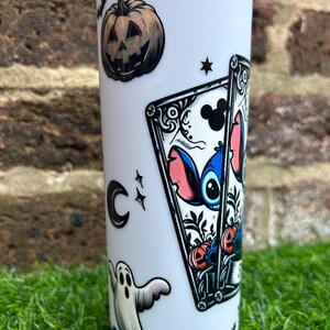Puede incluir: Un vaso blanco con tem&aacute;tica de Halloween con un personaje azul Stitch de Lilo & Stitch, una calabaza de Halloween, un fantasma y una carta de juego con una silueta de Mickey Mouse.