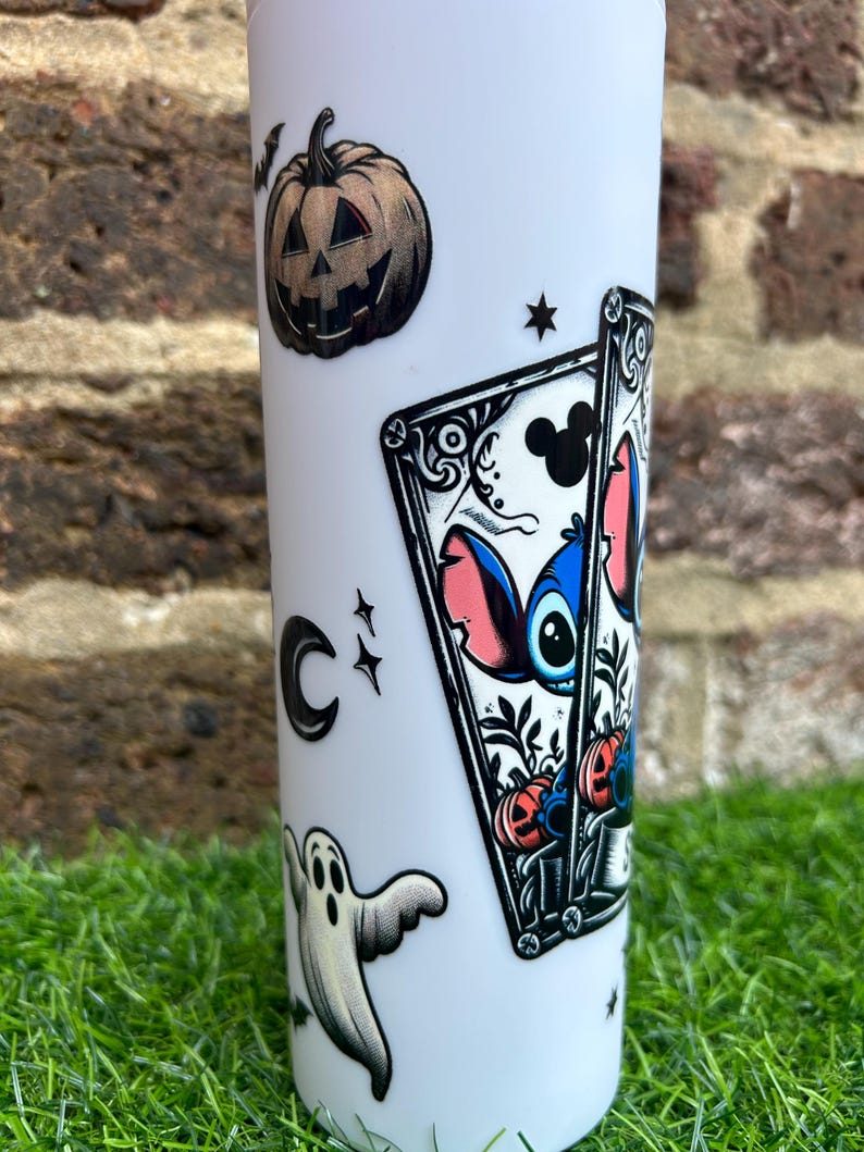Puede incluir: Un vaso blanco con tem&aacute;tica de Halloween con un personaje azul Stitch de Lilo & Stitch, una calabaza de Halloween, un fantasma y una carta de juego con una silueta de Mickey Mouse.