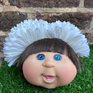 May include: A Cabbage Patch Kids doll head with brown hair and blue eyes, wearing a white flower crown. The doll's face is smiling, with rosy cheeks and two visible teeth. The doll is resting on green grass.