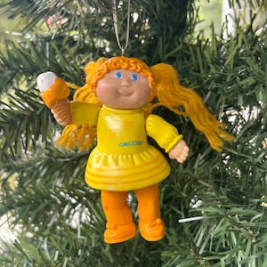 May include: A Cabbage Patch Kids Christmas tree ornament. The doll is wearing a yellow dress and orange shoes and holding an ice cream cone. The doll has yellow hair and blue eyes. The ornament is hanging on a green Christmas tree.