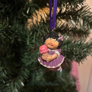May include: A vintage Cabbage Patch Kids Christmas tree ornament. The ornament depicts a doll holding an ice cream cone. The doll has black curly hair, blue eyes, and wears a purple dress. A purple ribbon is attached for hanging.