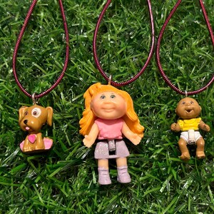May include: Three pendant necklaces with miniature figure charms. One is a brown dog with a pink heart, the second is a doll with blonde hair and a pink top, and the third is a baby doll in a yellow top.