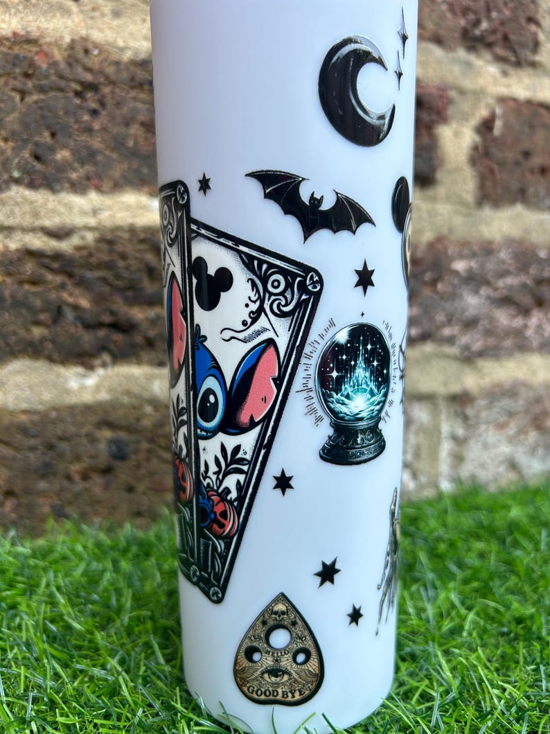 Puede incluir: Un vaso blanco con una variedad de pegatinas en blanco y negro. Las pegatinas incluyen una luna creciente, un murci&eacute;lago, una bola de cristal, una carta de tarot con un personaje de dibujos animados azul y rosa, y una tabla de Ouija negra y dorada con el texto "GOODBYE".