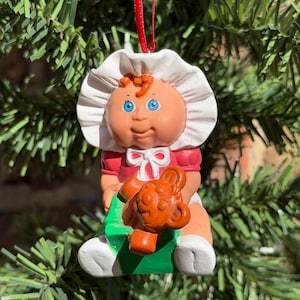 May include: A hanging ornament featuring a doll with a white bonnet, a red shirt with a white bow, and blue eyes, sitting on a green rocking horse while holding a brown teddy bear. The figure is suspended by a thin red ribbon.