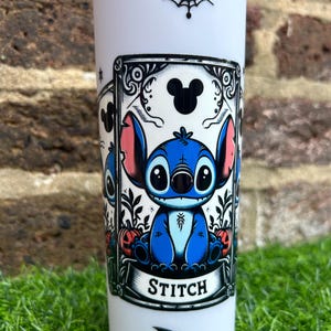 Puede incluir: Un vaso blanco con un dise&ntilde;o de telara&ntilde;a en blanco y negro en la parte superior. El dise&ntilde;o principal presenta un personaje azul de Stitch de Lilo & Stitch en un marco negro con la palabra "STITCH" en la parte inferior. Hay un murci&eacute;lago negro y dos estrellas en la parte inferior del vaso.