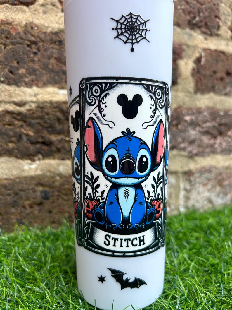 Puede incluir: Un vaso blanco con un dise&ntilde;o de telara&ntilde;a en blanco y negro en la parte superior. El dise&ntilde;o principal presenta un personaje azul de Stitch de Lilo & Stitch en un marco negro con la palabra "STITCH" en la parte inferior. Hay un murci&eacute;lago negro y dos estrellas en la parte inferior del vaso.