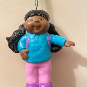 Puede incluir: Adorno de muñeca Cabbage Patch Kids. La muñeca tiene piel oscura, pelo negro en coletas y lleva una camisa azul con "CPK", pantalones rosas y zapatos morados. El adorno cuelga de una cuerda verde.