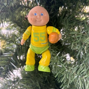 May include: A vintage Christmas tree ornament in the shape of a doll wearing a yellow football uniform with the number 55. The doll has blue eyes and is holding a brown football. The ornament is suspended from a Christmas tree.