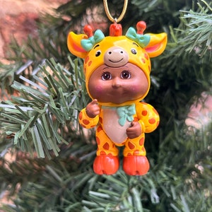 May include: A Christmas tree ornament depicting a figurine in a giraffe costume. The yellow outfit has brown spots, orange feet, and a green bow. The ornament hangs from a gold chain. The figurine is approximately 10 cm tall.