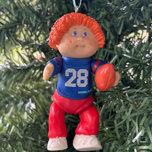 May include: A Cabbage Patch Kids Christmas ornament of a football player. The doll has orange hair, blue eyes, and is wearing a blue jersey with the number 28, red trousers, and white shoes. The ornament is holding an orange football.