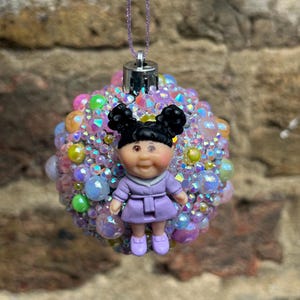 May include: A Christmas bauble with a doll figure in a lavender dress. The bauble is covered in iridescent beads and gems in pink, purple, and green hues. It hangs from a sparkly purple string.