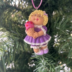 May include: A Cabbage Patch Kids Christmas tree decoration. The doll wears a purple dress and holds a pink ice cream cone. The ornament has a pink hanging string and is set against a backdrop of green Christmas tree branches.