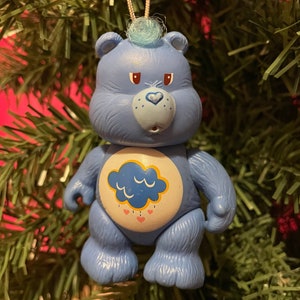Puede incluir: Adorno navideño azul de Care Bear, con un diseño de nube y lluvia en el vientre. El oso tiene un mechón de pelo azul claro y cuelga de un hilo dorado. El adorno está sobre un fondo de agujas de pino verdes.