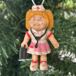 May include: A vintage Christmas ornament of a nurse doll. The doll has blonde hair, blue eyes, and wears a white uniform with a pink skirt and red accents. A black stethoscope hangs around the neck, and the doll carries a black medical bag. The ornament hangs from a silver string.