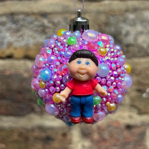 May include: A decorative Christmas ornament featuring a doll figure in a red shirt and blue pants. The ornament is covered in iridescent pink and purple beads and gems, with accents of yellow and green. The doll has brown hair and blue eyes.