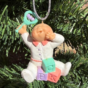May include: A hanging ornament featuring a baby figurine wearing a white outfit with a pink collar. The baby holds a light green rattle and sits on three stacked blocks coloured purple, green, and orange, which are embossed with the letters P, G, and K.