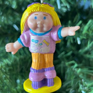 May include: A Cabbage Patch Kids ornament with a yellow base. The doll has yellow hair, blue eyes, and is wearing a pink shirt with the words "THEM" and "BY" on it. The doll is wearing orange trousers and purple shoes.