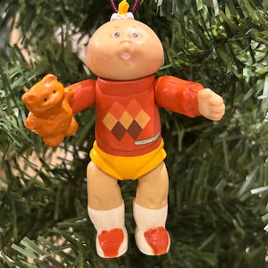 May include: A Cabbage Patch Kid ornament wearing a red and yellow sweater, yellow trousers, and white socks. The ornament is holding a brown teddy bear. The ornament is hanging from a red string.