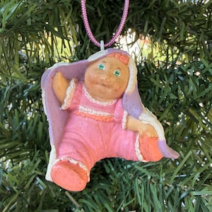 May include: A hanging ornament featuring a small doll with a round face, green eyes, and orange hair, wearing a pink outfit with white trim. The doll is partially wrapped in a light purple blanket with white edges and hangs from a pink cord.