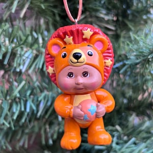 May include: A small, orange figurine of a baby wearing a lion costume, holding a ball with blue star designs. The figurine has a red mane with yellow stars and is suspended by a pink string, suitable for use as an ornament.