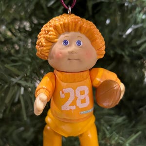 May include: Orange and white Cabbage Patch Kid ornament wearing a football jersey with the number 28. The ornament is holding a brown football.