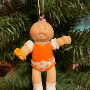 May include: A Cabbage Patch Kids Christmas tree ornament. The doll has a bald head, rosy cheeks, and is wearing an orange outfit with a white nappy featuring carrot designs. It holds a yellow spoon and white socks. The ornament hangs from a white string.