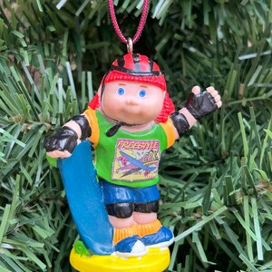 May include: A Christmas tree ornament depicting a skater doll with red hair, wearing a red helmet, green shirt with "FREESTYLE CPK" text, blue shorts, and black protective gear. The doll holds a blue skateboard and stands on a yellow base.