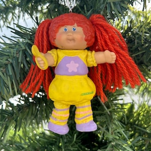 May include: A Cabbage Patch Kids Christmas tree decoration. The doll has red yarn pigtails, blue eyes, and a yellow outfit with a purple top and a pink star. The doll is holding a yellow accessory. The ornament hangs from a silver chain.