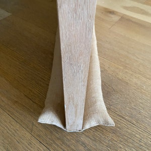 May include: A close-up of a doorstop made of natural burlap fabric. The doorstop is placed at the bottom of a wooden door frame.