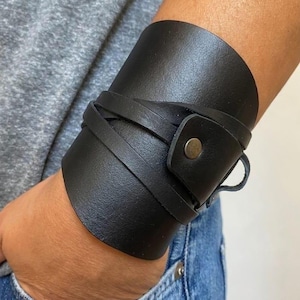 Op de afbeelding: Een brede zwarte leren manchetarmband met een glad oppervlak en een wikkelbandontwerp. De armband wordt vastgezet met een enkele metalen drukknoopsluiting, wat zorgt voor een gelaagd uiterlijk dat de pols volledig omsluit.