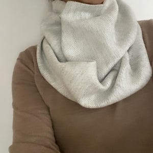 May include: A white infinity scarf with a textured knit pattern. The scarf is folded and draped around the neck.