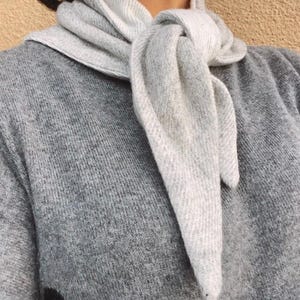 Könnte beinhalten: Ein hellgrauer Strickschal, der einfach um den Hals geknotet ist und über einem passenden hellgrauen Strickpullover getragen wird. Der Schal besteht aus einem weichen, strukturierten Stoff mit einem dezenten Rippmuster für ein einheitliches Bild.