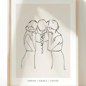 May include: A framed line art print depicting three figures in a minimalist style. The artwork uses black lines on a beige background, with the names "SARAH | GRACE | CHLOE" printed at the bottom. The frame is made of light-coloured wood.