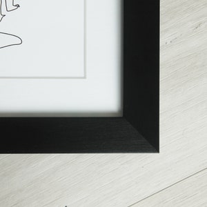 K&ouml;nnte beinhalten: Schwarz gerahmter Kunstdruck mit einer minimalistischen Strichzeichnung auf wei&szlig;em Hintergrund. Das Kunstwerk ist in einem schwarzen Holzrahmen gerahmt und wird auf einer hellen Holzoberfl&auml;che pr&auml;sentiert.