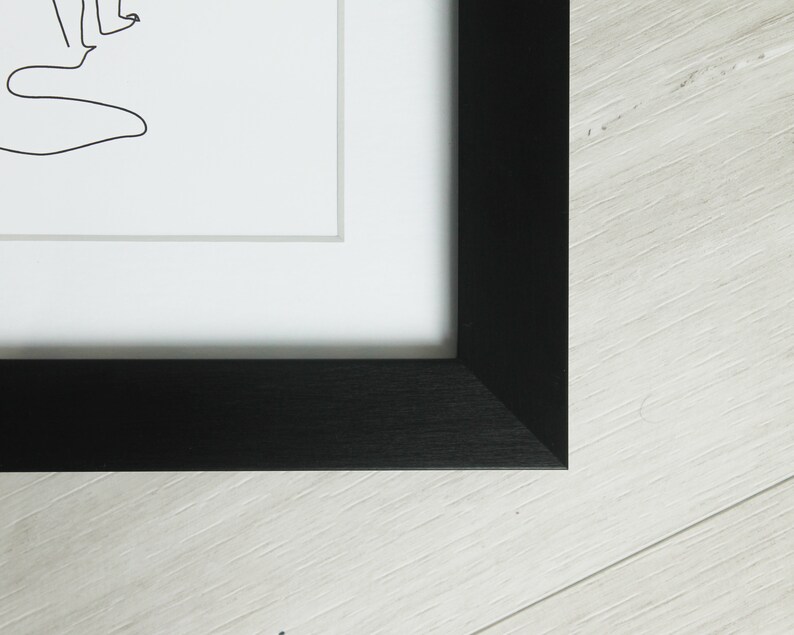 K&ouml;nnte beinhalten: Schwarz gerahmter Kunstdruck mit einer minimalistischen Strichzeichnung auf wei&szlig;em Hintergrund. Das Kunstwerk ist in einem schwarzen Holzrahmen gerahmt und wird auf einer hellen Holzoberfl&auml;che pr&auml;sentiert.