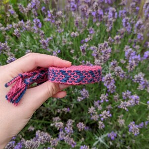 May include: A close-up of a pink and blue woven bracelet with a fringe on one end. The bracelet is held in a hand against a background of purple lavender flowers.