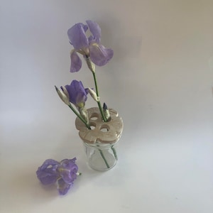 May include: A white ceramic flower frog with multiple holes sits atop a clear glass vase. Two purple irises are arranged in the vase.