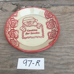May include: A white ceramic plate with red trim and a red design of Santa Claus holding a sign that says "Cookies for Santa".