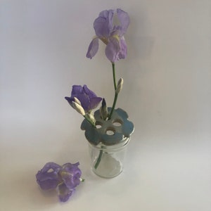 May include: A blue ceramic flower frog sits atop a clear glass jar with a single purple iris flower and buds. The flower frog has six holes for stems.
