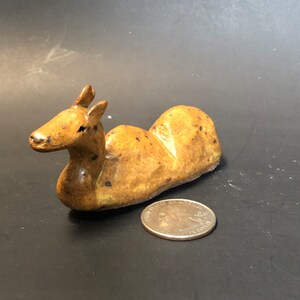 May include: A small ceramic sculpture of a brown deer lying down. The deer has a speckled brown glaze and is about the size of a quarter.