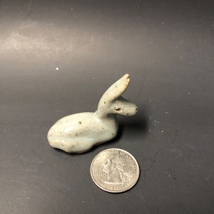 May include: A small, gray ceramic sculpture of a rabbit. The rabbit is lying down with its head turned to the side. The sculpture is about the size of a quarter.