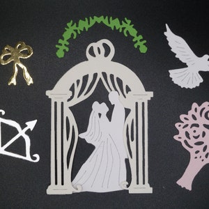 May include: White paper cutouts of a wedding arch, a bride and groom, a dove, a bow, an arrow, and a bouquet of roses. The arch is decorated with a green paper cutout of a leafy garland.