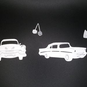 Puede incluir: Un conjunto de formas de papel blanco troqueladas que incluyen dos autom&oacute;viles cl&aacute;sicos al estilo de los a&ntilde;os 50, dos flechas direccionales con bordes punteados y un par de dados colgantes sobre un fondo negro s&oacute;lido.
