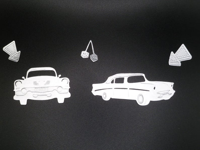 Puede incluir: Un conjunto de formas de papel blanco troqueladas que incluyen dos autom&oacute;viles cl&aacute;sicos al estilo de los a&ntilde;os 50, dos flechas direccionales con bordes punteados y un par de dados colgantes sobre un fondo negro s&oacute;lido.