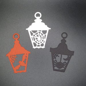 May include: Three Halloween-themed paper lanterns, one white, one orange, and one black. Each lantern has a different design, including a spider web, a bat, and a spider.