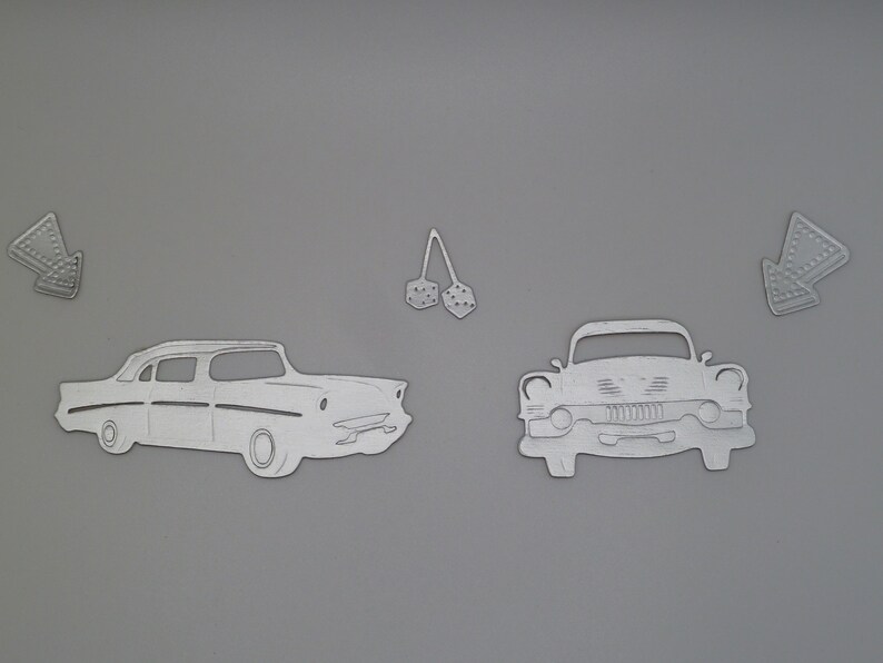 Puede incluir: Un conjunto de formas met&aacute;licas troqueladas de color plateado que incluyen dos coches cl&aacute;sicos, un par de dados colgantes y dos se&ntilde;ales de flecha. Las formas son planas con contornos y detalles en relieve sobre un fondo gris s&oacute;lido.