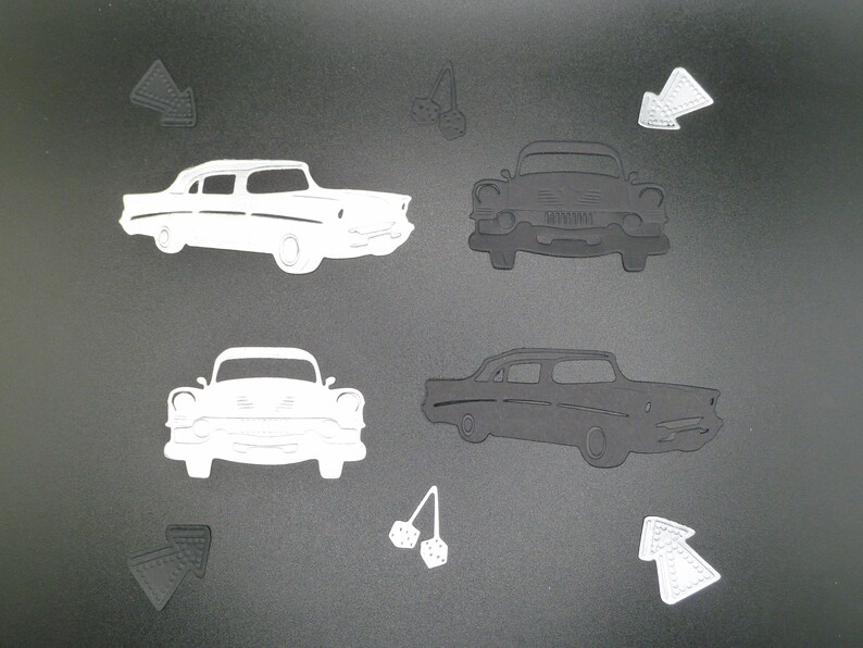 Puede incluir: Una colecci&oacute;n de formas troqueladas que incluye dos siluetas de coches cl&aacute;sicos en blanco y dos en negro, dos flechas en blanco y dos en negro, y dos pares de dados colgantes en blanco y negro sobre una superficie gris oscuro.
