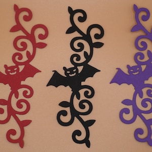 May include: Three paper cutouts of bats hanging from a vine. The bats are red, black, and purple.