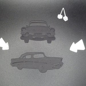 Puede incluir: Dos siluetas negras troqueladas de un coche cl&aacute;sico, una con vista frontal y otra con perfil lateral. El conjunto incluye dos formas blancas troqueladas, una que representa un par de dados colgantes y dos acentos blancos en forma de flecha.