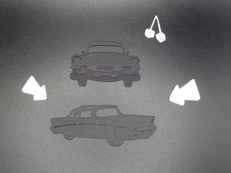 Puede incluir: Dos siluetas negras troqueladas de un coche cl&aacute;sico, una con vista frontal y otra con perfil lateral. El conjunto incluye dos formas blancas troqueladas, una que representa un par de dados colgantes y dos acentos blancos en forma de flecha.